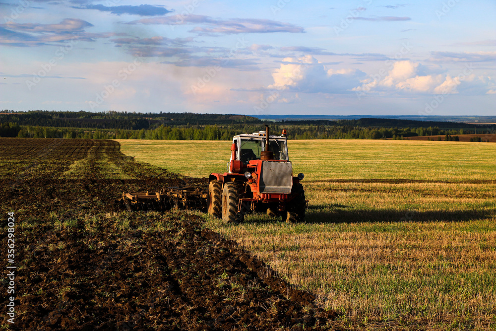 Obraz premium tractor plows and harrows land in large field on sunny spring day. preparing soil for planting crops, plowing soil with tractor with disk plow.