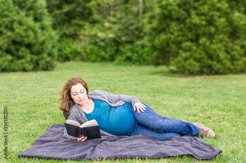 pregnant woman is reading a book on a green field in a park. © olegmayorov