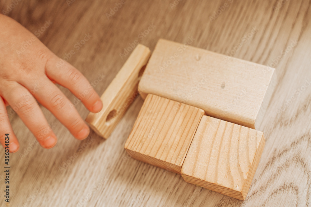 in the foreground the child's hands and he is building a house out of wooden beige cubes in the background a wooden beige floor