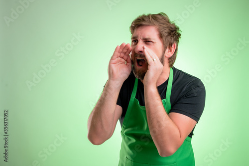 Supermarket employee with green apron and black t-shirt shouting with his hand to his mouth