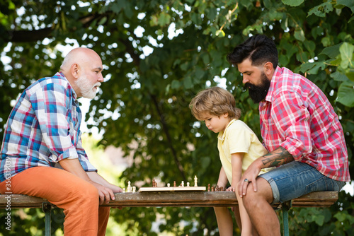 Boy with father and grandfather. Little boy playing chess with his Grandfather and Father. Summer and active holidays. Senior man thinking about his next move in a game of chess. © Volodymyr