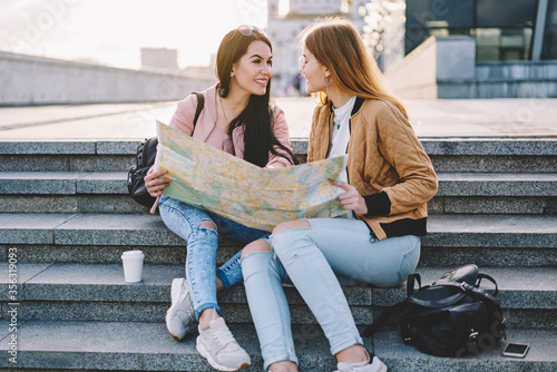 Two hipster girls discussing route of future travel sitting outdoors in urban setting