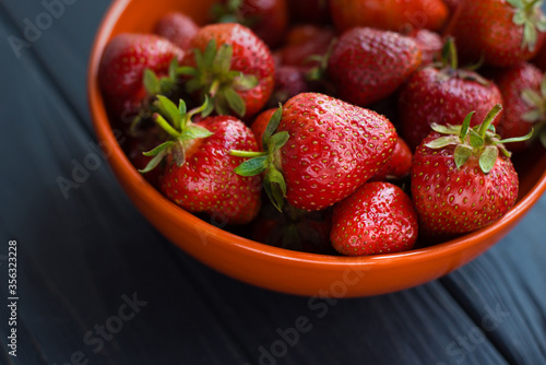 Close-up fresh ripe delicious strawberries in a orange bowl plate on wooden black table background. Antioxidant nutrition, healthy organic food concept. Copyspace. Flat lay. Daylight
