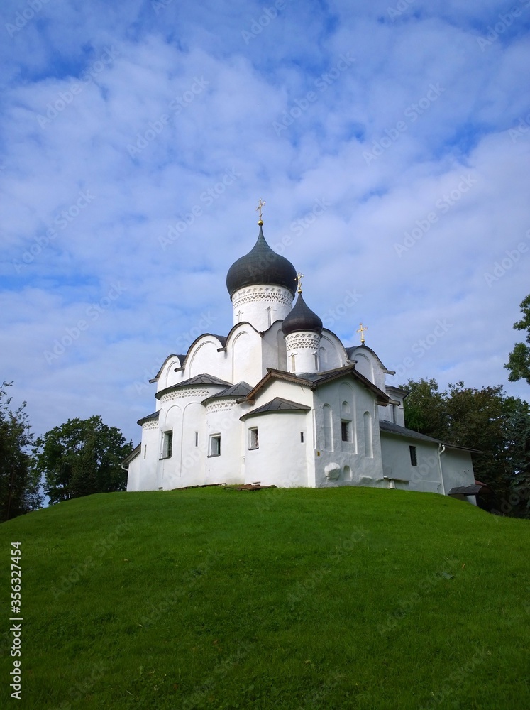 Ancient orthodox christian stone temple. Pskov, Russia. The Church of St. Basil the Great on the Hill on a background of green grass and beautiful blue sky with white clouds. Unique architecture. Ancient orthodox christian stone temple. Pskov, Russia. The Church of St. Basil the Great on the Hill on a background of green grass and beautiful blue sky with white clouds. Unique architecture.