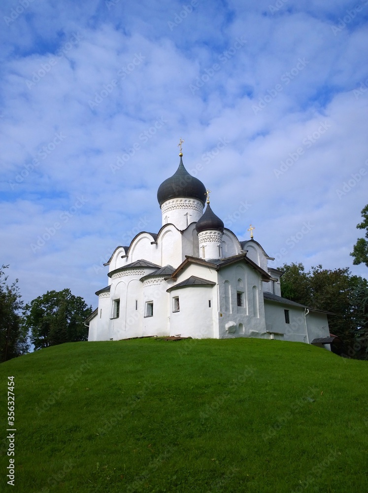 Ancient orthodox christian stone temple. Pskov, Russia. The Church of St. Basil the Great on the Hill on a background of green grass and beautiful blue sky with white clouds. Unique architecture.