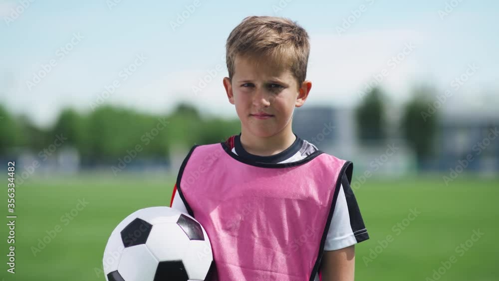 Portrait of a football player boy, a young boy stands near a football field and looks at the camera, holding a football in his hands, blurred background.