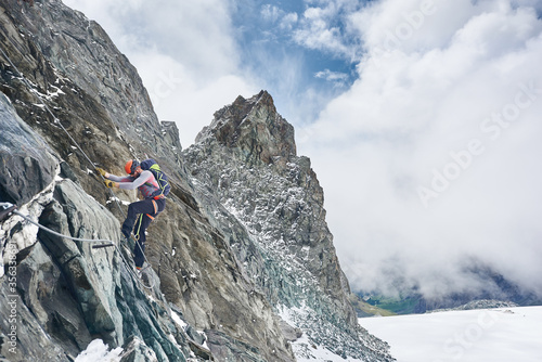 Rock climber in safety helmet using fixed rope while ascending rocky mountain. Male alpinist with backpack climbing natural rock formation. Concept of alpinism and winter mountaineering.