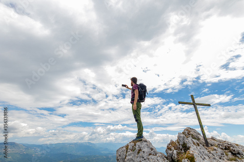 Man takes a photo on top of a mountain with a cross. Trekking and hiking in the mountains in summer. Mountain tourism