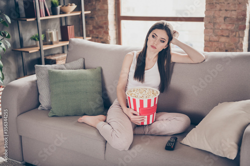 Full length photo of tired bored girl sit divan watch uninteresting movie hold pop corn box in living room house indoors