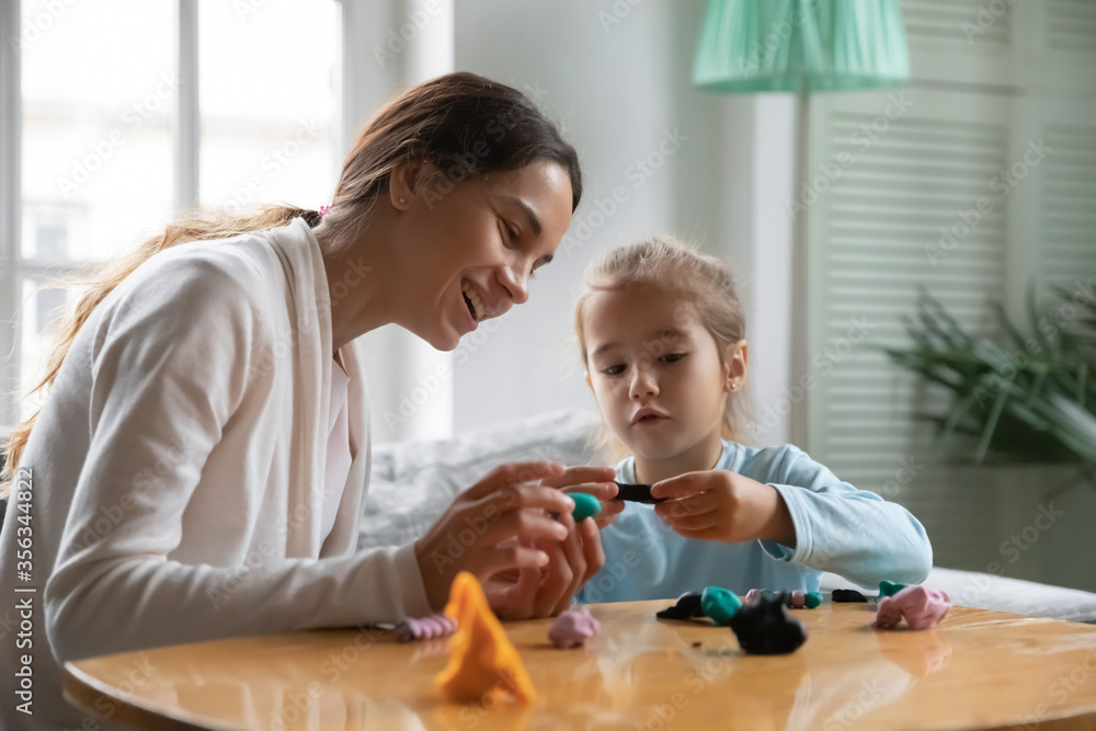 Smiling young mom sit at table play with plasticine modeling clay with little daughter, happy Caucasian mother or nanny have fun engaged in creative activity with Play-Doh with small girl child