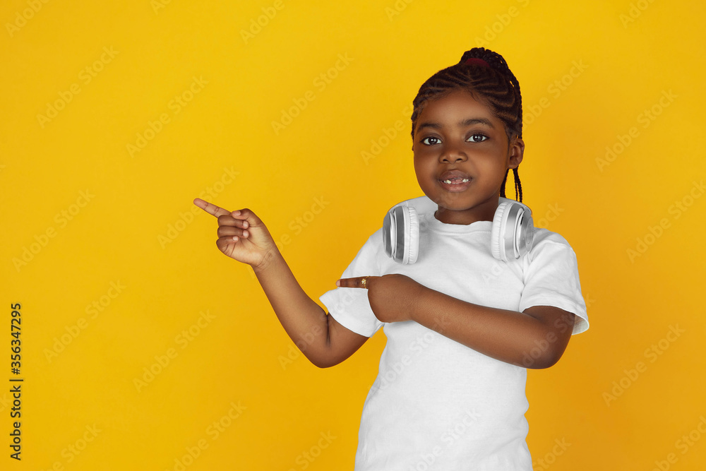 Pointing on, putting on headphones. Little african-american girl's portrait on yellow studio background. Cheerful kid. Concept of human emotions, facial expression, sales, ad. Copyspace. Looks cute. Pointing on, putting on headphones. Little african-american girl's portrait on yellow studio background. Cheerful kid. Concept of human emotions, facial expression, sales, ad. Copyspace. Looks cute.