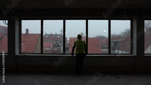 Lonely tired construction worker old man with a vest and helmet looks out the window on a construction site centered in a large window looking out on a rainy moody day silhouette and over his shoulder photo