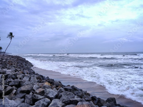 A picture of beach with rocks along with a coconut tree on the shore and cloudy sky on background. The sea is violent with strong waves in this picture. Kappil Beach (Varkala Town), Kerala. photo