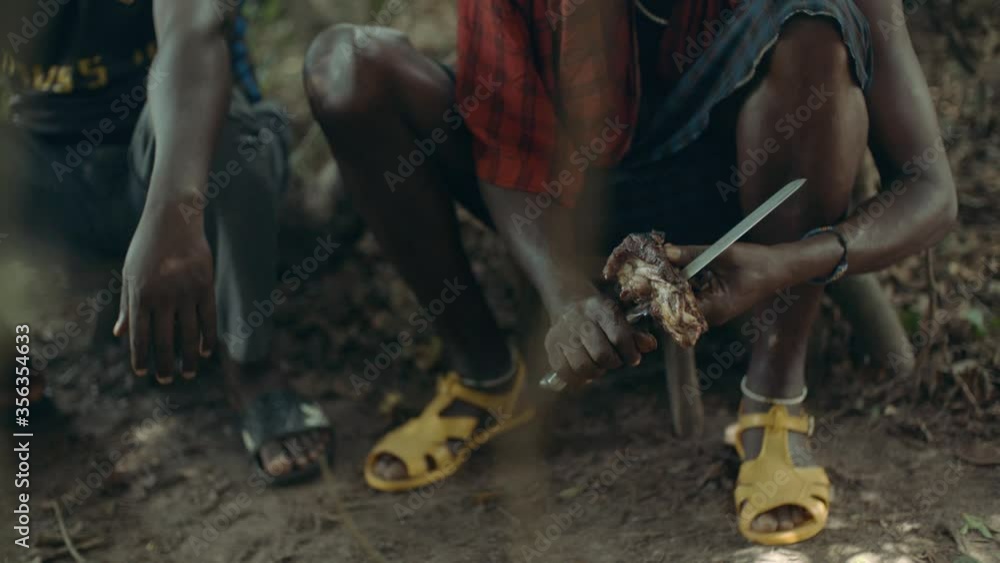 An African sitting in a squat and wearing traditional Maasai clothes is cutting a piece of meat with a knive. We can not see their faces but the dancing sunlight on the ground. An African sitting in a squat and wearing traditional Maasai clothes is cutting a piece of meat with a knive. We can not see their faces but the dancing sunlight on the ground.