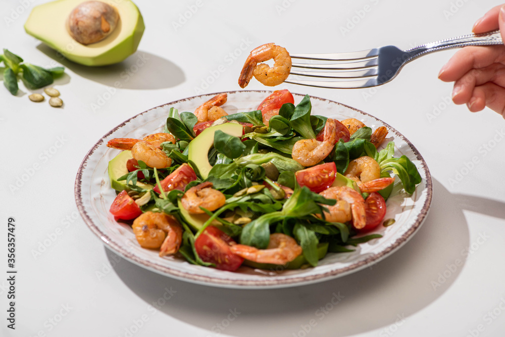 partial view of woman eating fresh green salad with cherry tomatoes, shrimps and avocado on white background