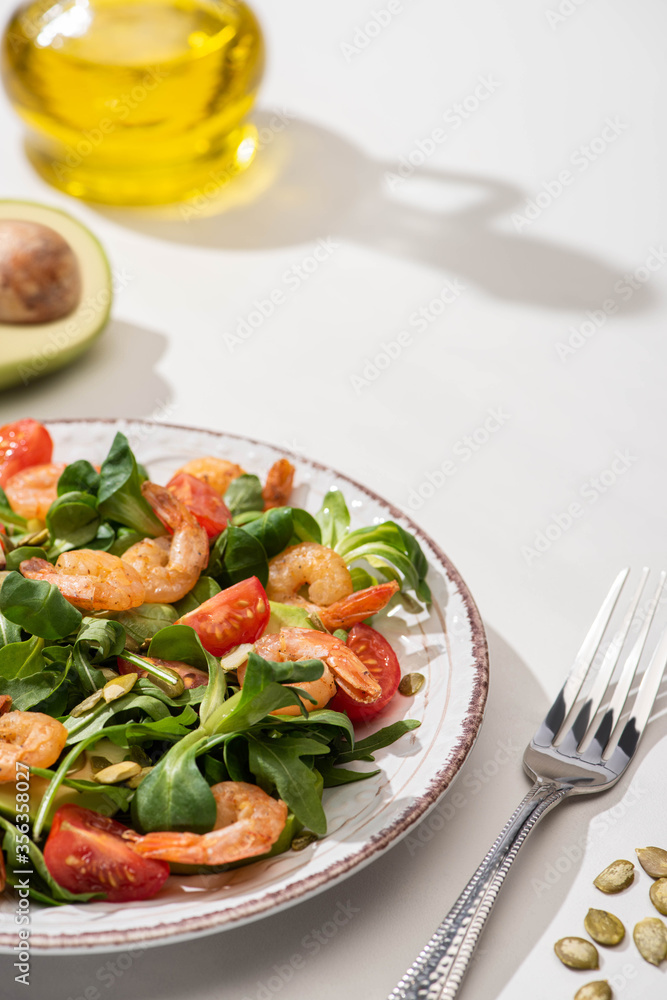 selective focus of fresh green salad with shrimps and avocado on plate near fork and ingredients on white background selective focus of fresh green salad with shrimps and avocado on plate near fork and ingredients on white background