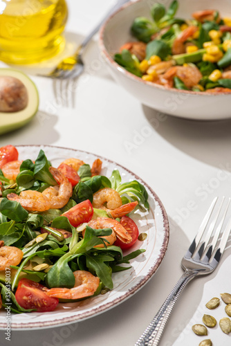 selective focus of fresh green salad with shrimps and avocado on plates near forks on white background