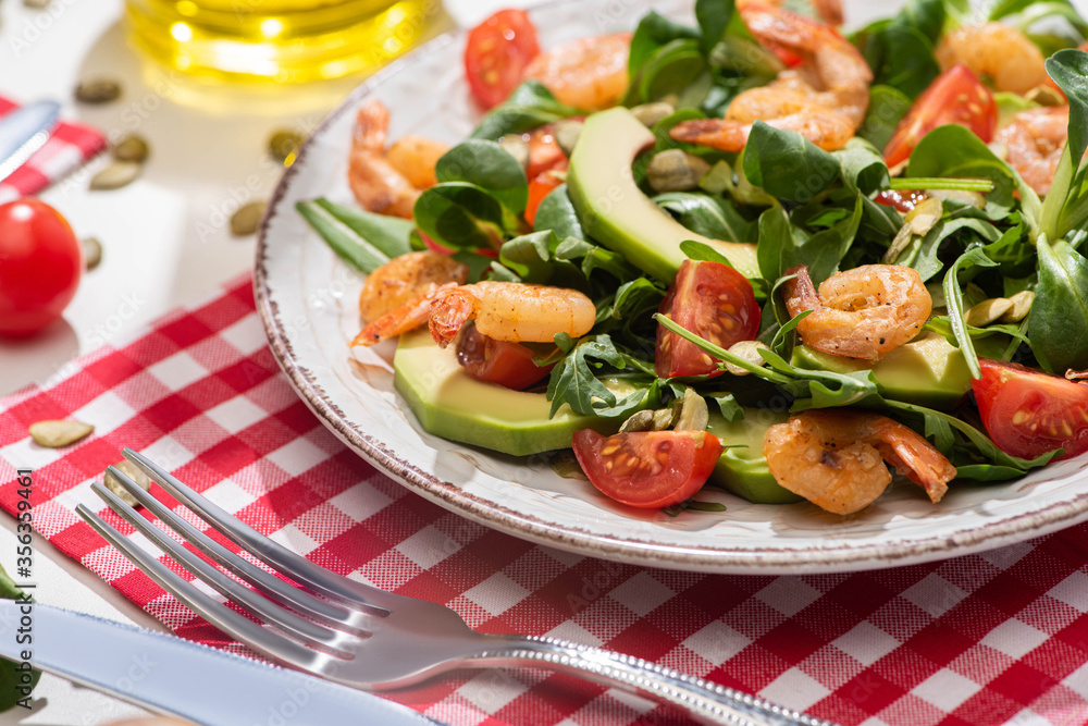 selective focus of fresh green salad with shrimps and avocado on plate near cutlery on plaid napkin selective focus of fresh green salad with shrimps and avocado on plate near cutlery on plaid napkin