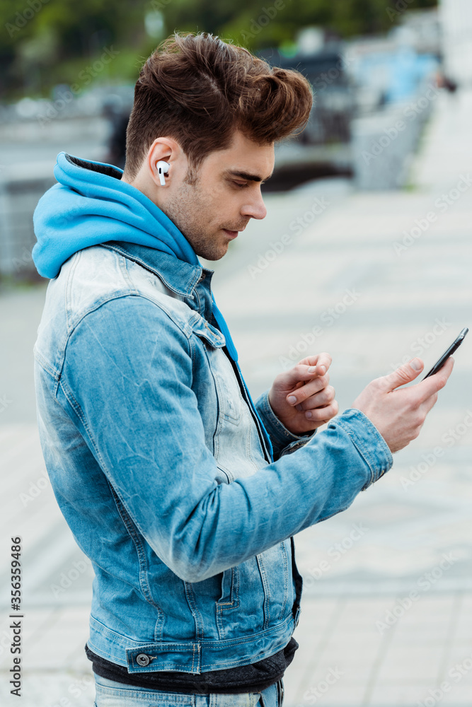 Side view of handsome young man using earphones and smartphone on urban street
