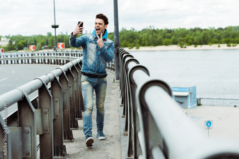 Selective focus of cheerful man showing peace sign while taking selfie with smartphone on bridge