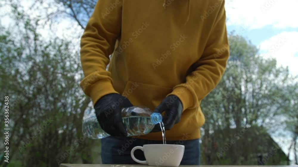 person in yellow hoodie and black sterile gloves pours water into white cup and drinks standing against village house slow motion person in yellow hoodie and black sterile gloves pours water into white cup and drinks standing against village house slow motion