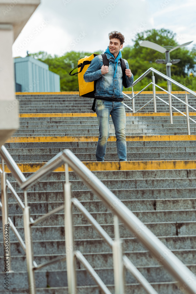 Selective focus of smiling courier with thermal backpack walking on stairs with railing Selective focus of smiling courier with thermal backpack walking on stairs with railing