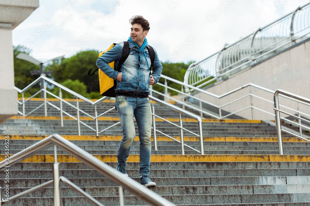 Selective focus of smiling courier with thermo backpack walking down on stairs with railing
