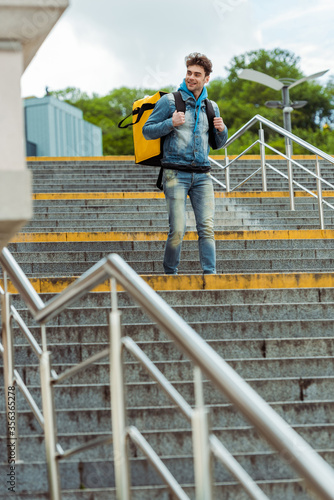 Selective focus of smiling courier with thermal backpack walking on stairs with railing