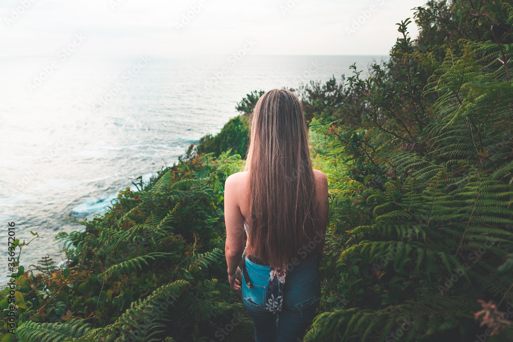 Young girl with white shirth and blue jeans walking through the forest next to the sea.