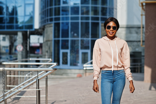 Hipster african american girl wearing pink hoodie,sunglasses and jeans posing at street against office building with blue windows.