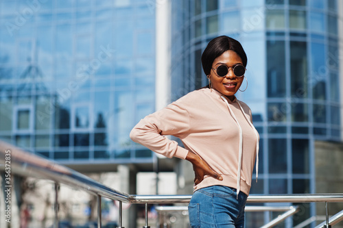 Hipster african american girl wearing pink hoodie,sunglasses and jeans posing at street against office building with blue windows.