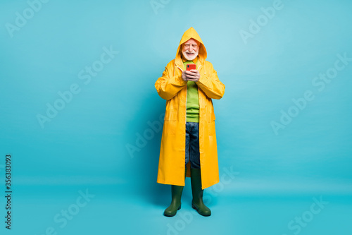 Full length body size view of his he nice cheerful cheery grey-haired man wearing long yellow topcoat using cell chatting free time isolated on bright vivid shine vibrant blue color background