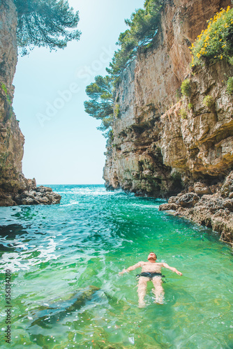 young man swimming on back in sea at bay between cliffs
