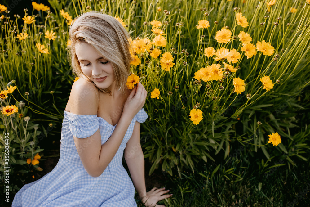 Young woman with long blonde hair, wearing country style dress, sitting in a garden with yellow flowers in golden hour. Young woman with long blonde hair, wearing country style dress, sitting in a garden with yellow flowers in golden hour.
