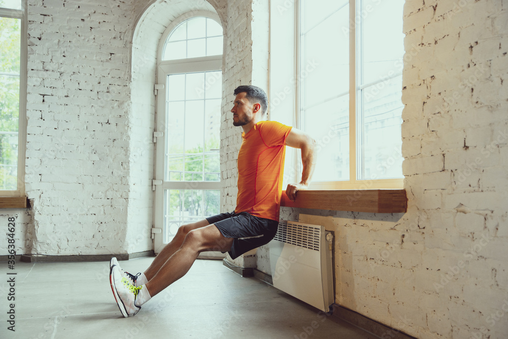 Fototapeta premium Sit ups. Young caucasian man training at home during quarantine of coronavirus outbreak, doing exercises of fitness, aerobic. Staying sportive during insulation. Wellness, sport, movement concept.