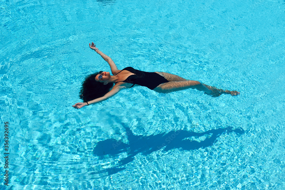 Young beautiful sexy slim tanned caucasian woman with brunette hair in black swimwear relaxing and lying in blue water of swimming pool at tropical resort. Vacation concept. Top poinit of view