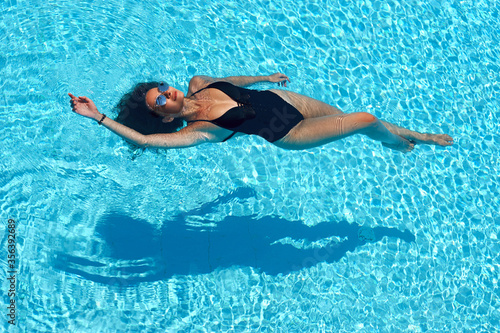 Young beautiful sexy slim tanned caucasian woman with brunette hair in black swimwear relaxing and lying in blue water of swimming pool at tropical resort. Vacation concept. Top poinit of view