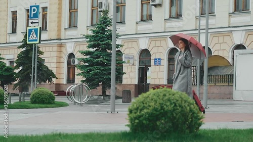 A young girl in a gray travel coat walks near an empty train station with a suitcase under an umbrella. Slow motion