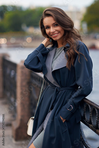 Happy joyous young woman standing and posing at river embankment in city. Pretty girl in dress and raincoat with makeup and wavy hair. Brunette caucasian female.