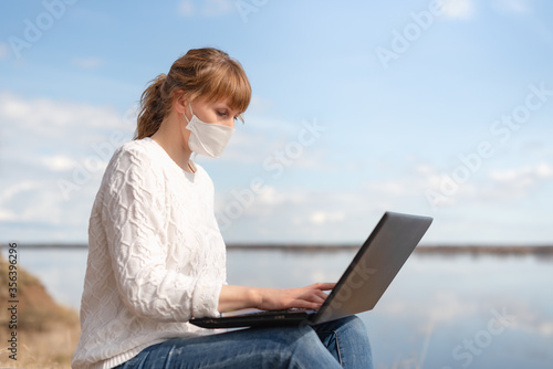 masked woman working remotely on a laptop outdoors, on the background of the river and sky