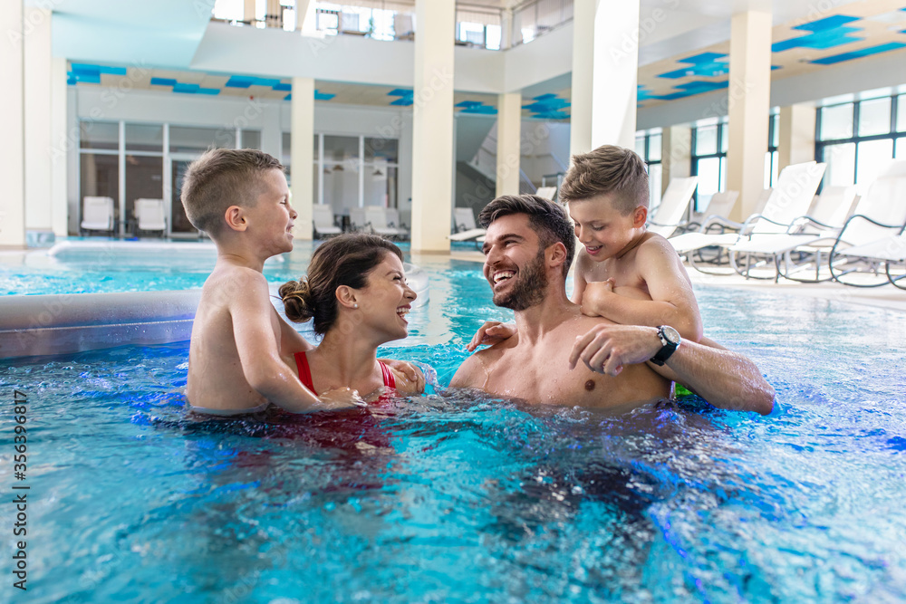 Smiling family of four having fun and relaxing in indoor swimming pool at hotel resort. - obrazy, fototapety, plakaty 