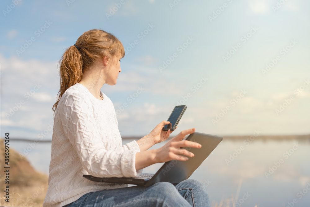 a woman in a white sweater sits in nature and works remotely on a laptop, looks at the phone.
