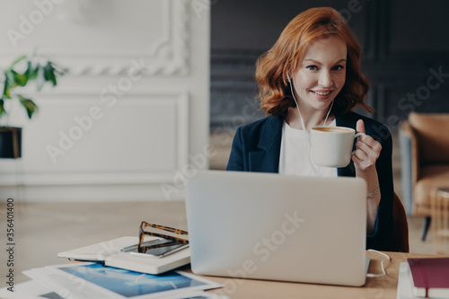 Happy young redhead woman watches training webinar on laptop computer, browses information for project, drinks coffee or espresso, poses at desktop, uses earphones for e learning, autodidact