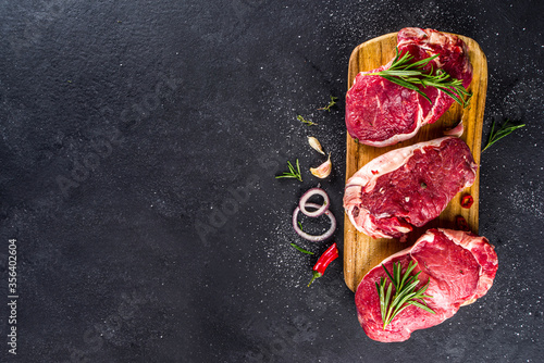 Raw meat, beef steak on cutting board with rosemary and spices, black background, top view copy space