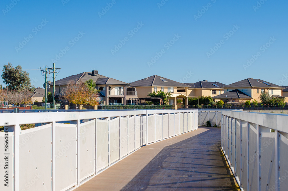 A pedestrian boardwalk leads to a residential neighbourhood with some two-storey modern houses. Sanctuary Lakes, Point Cook, Melbourne, VIC Australia.
