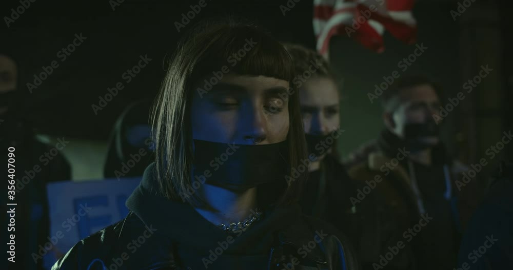 Close up view of young female protestor with tape on mouth looking to camera while standing at crowd . Demonstrators striking against racism and police brutality at night street Close up view of young female protestor with tape on mouth looking to camera while standing at crowd . Demonstrators striking against racism and police brutality at night street