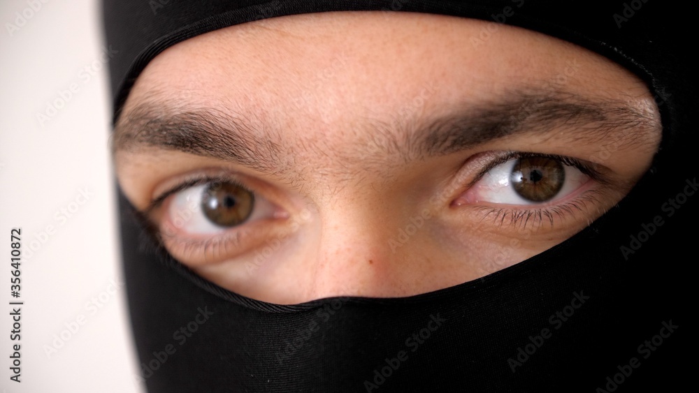 Close-up portrait of a man in a black balaclava. Brown eyes close up. Thief or burglar looks at the camera. Riots, robbery and marauding. Face of the criminal world threatens honest citizens. Close-up portrait of a man in a black balaclava. Brown eyes close up. Thief or burglar looks at the camera. Riots, robbery and marauding. Face of the criminal world threatens honest citizens.