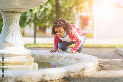 Cute little dark skinned hispanic girl playing with paper ship by a fountain on spring or autumn day. Active leisure for children. Child having fun outdoor. Sun glare effect.