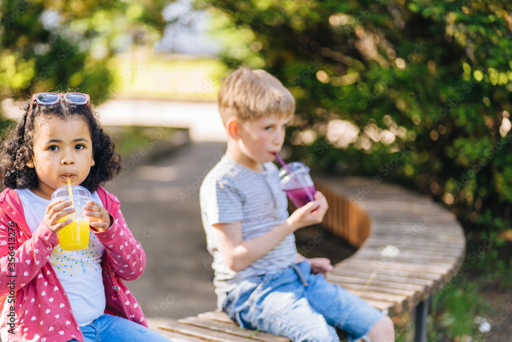 Children do not have racism. Little Hispanic Preschooler Girl and Cute White Caucasian Boy Sitting On The Bench, drinking a fruit smoothie in summer city park. Children do not have racism. Little Hispanic Preschooler Girl and Cute White Caucasian Boy Sitting On The Bench, drinking a fruit smoothie in summer city park.