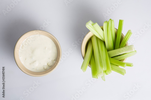 close up isolated flat lay top view shot of a bowl of crunchy juicy green celery sticks next to a white cup of blue cheese dipping sauce on a white background